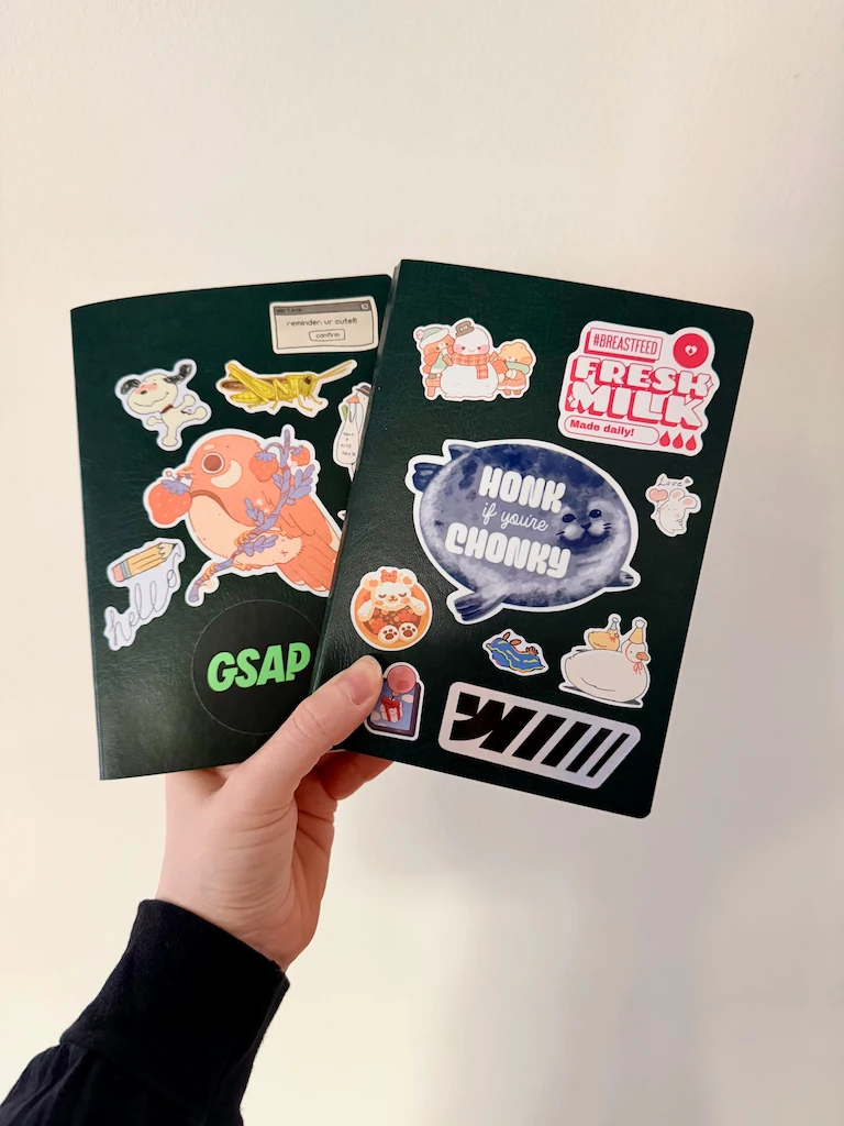 My hand holding two B6 journals with covers so dark green they look black. The covers are covered in cutesy stickers described after this amage.