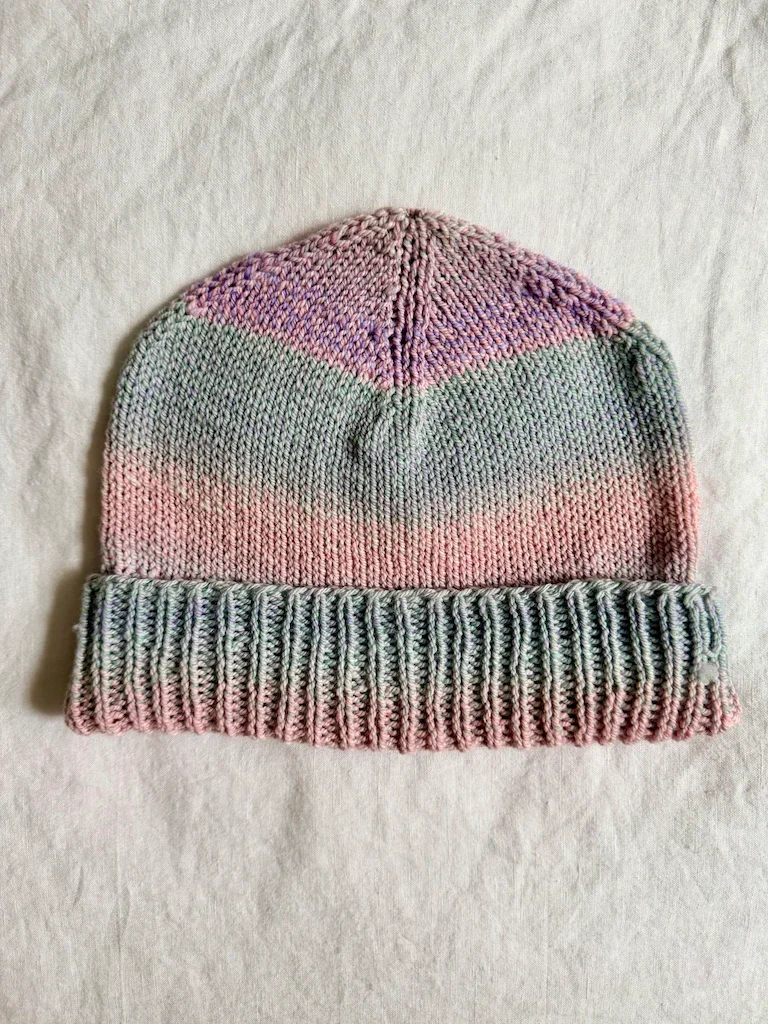 A knit beanie in a gradient of blue, pink, and purple yarn, with a ribbed brim rolled up on the bottom of the hat from the bottom