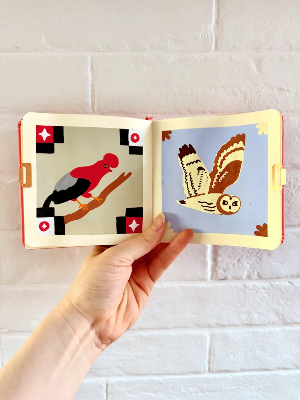 Left page: a Posca drawing of a red and black bird with a big red poof on its head. It's sitting on a thin branch. In the corners are red, black, and white rectangular motifs with sparkles on the inside of each corner. Right page: a cream and brown owl in flight. In the corners are brown and cream sort of feathery motifs.