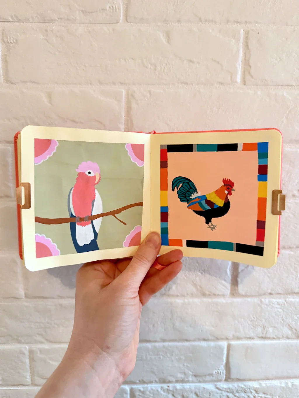 Left: a parrot-like bird in shades of pink, white, and grey. The corners have pink crenallated motifs that kind of look like fans. On the right is a rooster-looking bird of many colors. The page border is a series of bars in the colors of the bird; it kind of looks like LEGO bricks all lined up.