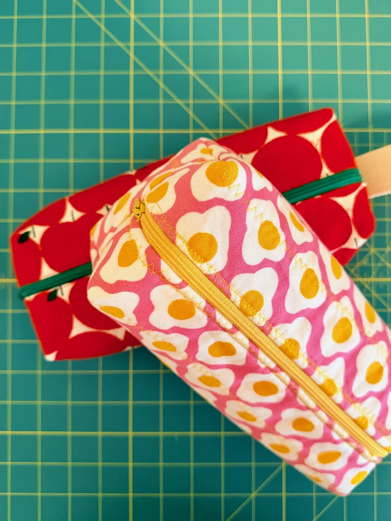 Two long, boxy fabric pouches piled on top of each other on a self-healing cutting mat. One pouch has pink, yellow, and white egg fabric and a yellow zipper. The bottom pouch has red, green, and white apple fabric with a dark green zipper.