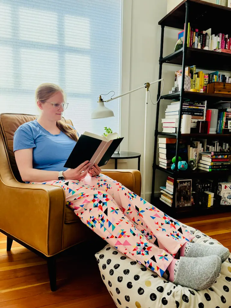 Me sitting in a chair pretending to read a book while my feet are propped up on a pouf. I am wearing pale pink PJ bottoms that have a pattern of scattered geometric quilt blocks on them.