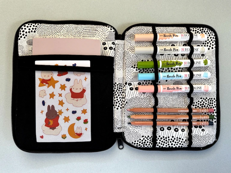 The case laid out flat and filled with materials: a notebook, Miffy stickers, Ecoline markers, and colored pencils