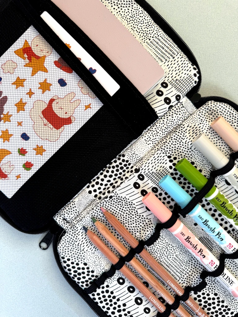 A jaunty close-up of a fabric supply case in black and white fabric, filled up with a notebook, Miffy stickers, Ecoline markers, and colored pencils