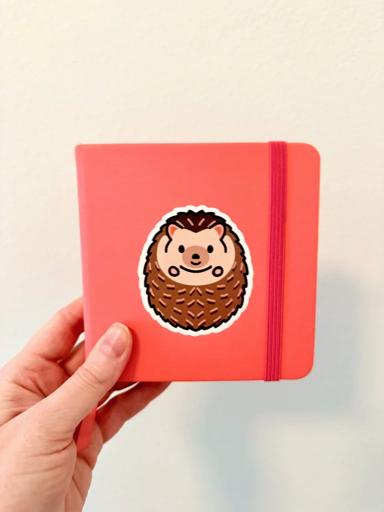 A small, square, coral sketchbook with a cute illustrated sticker of a hedgehog on it