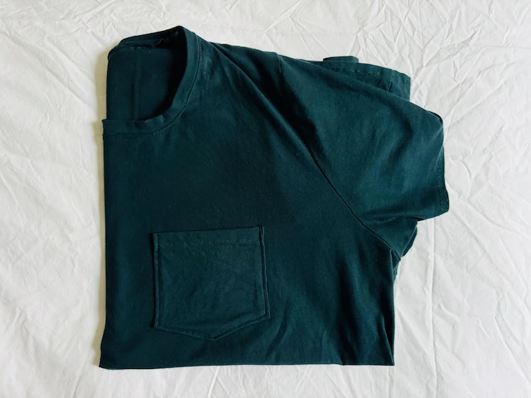 The t-shirt folded in quarters on top of a slightly wrinkly white fabric background. The pocket is on fairly straight and has two angled bottom sides.