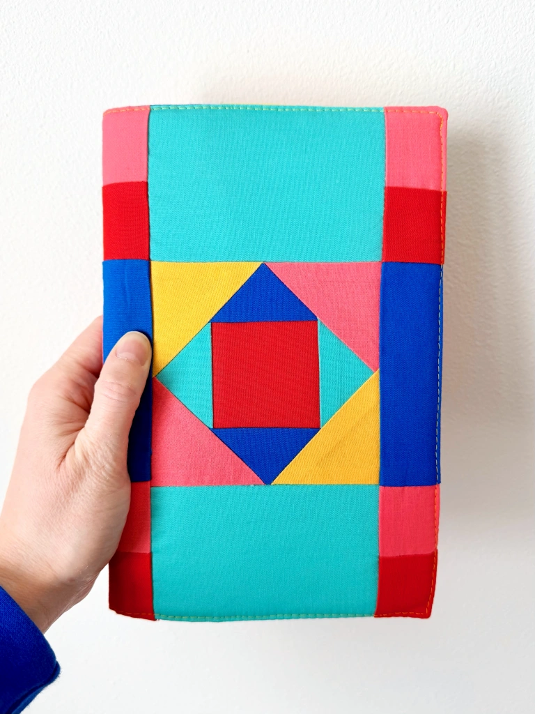 On the front, the central block has a red square surrounded by a diamond made of blue and teal triangles. The square around that is made from pink and yellow triangles. Around the central block are rectangles in teal, blue, pink, and red.