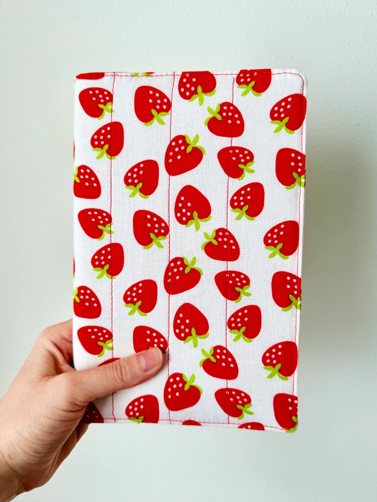 A quilted portrait-orientation sketchbook cover. The white fabric has bright, graphic strawberries in red and lime green on it. The quilting is done with red thread in vertical lines 1.5 inches apart