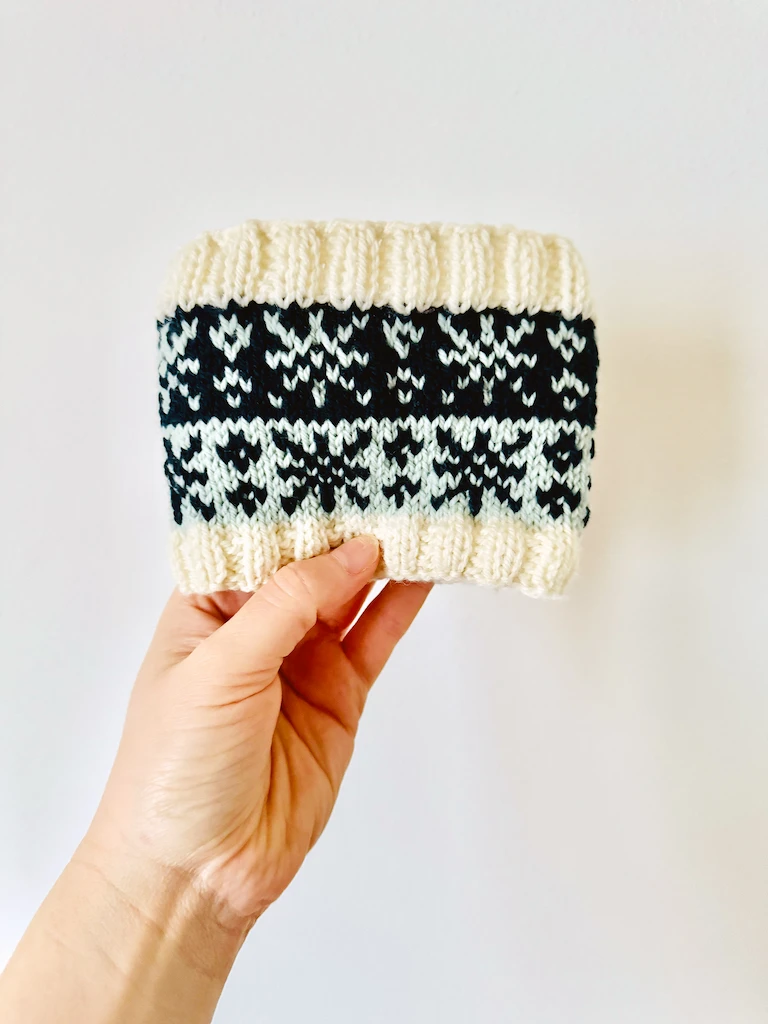 My hand holding a knitted headband that's been folded in half. There is cream ribbing on the lower and upper edge. Between these are two bands of color, one light blue and one dark blue. On each band is a simple knitted snowflake pattern in the contrasting color.