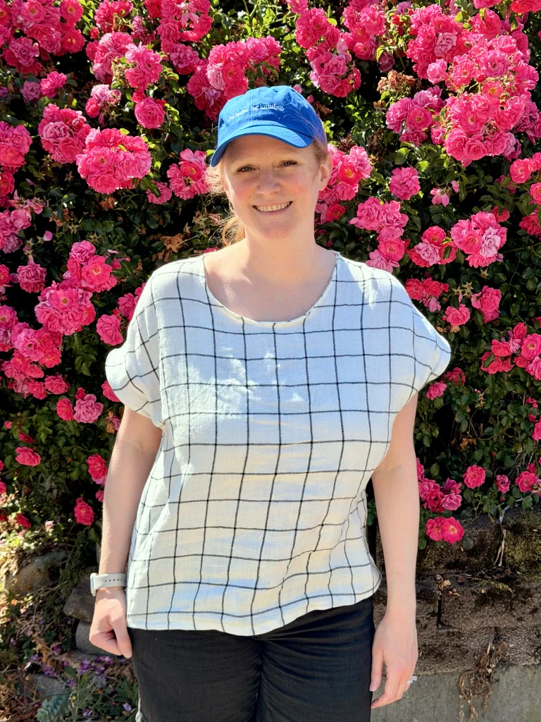 Me standing in front of a rose bush heavy with bright pink blossoms. I'm wearing a boxy short-sleeved shirt, sewn in white linen with a dark grey window-pane pattern. The pattern is on the larger side, vs very fine details.