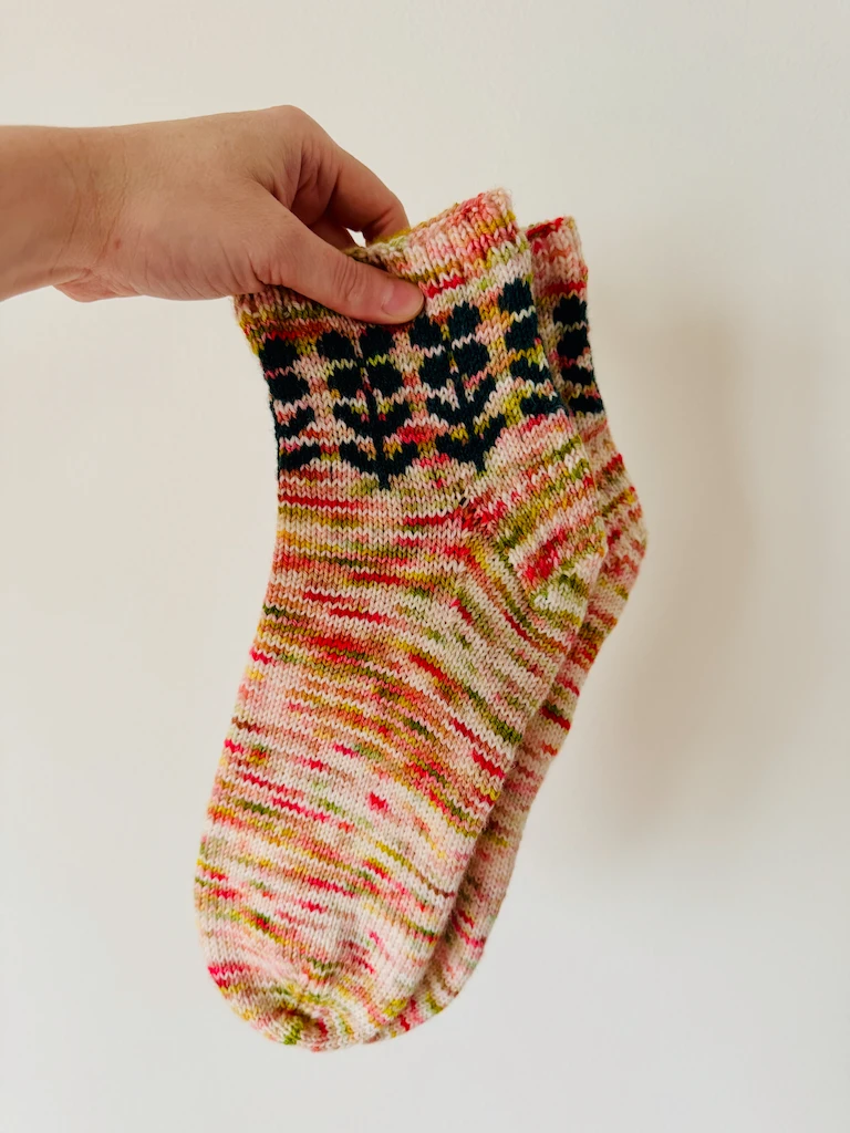 A pair of socks in variegated yarn of cream, pinks, and lime greens. Around the cuffs is a dark green flower motif.