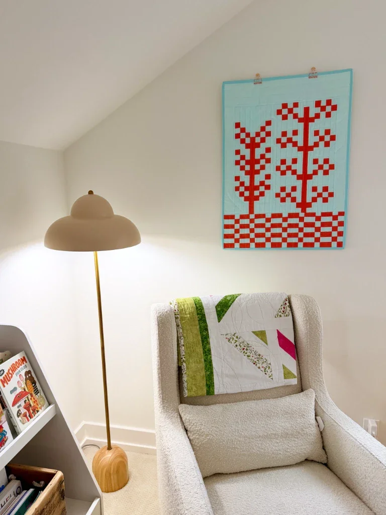 The quilt in context of a nursery with off-white walls. In the corner of a room you can see part of a white bookshelf, a nubbly off-white arm chair with a pink, green, and white quilt hanging over the back, and an off-white lamp that's shaped sort of like a boob. Hanging above the chair and next to the lamp is my new quilt.