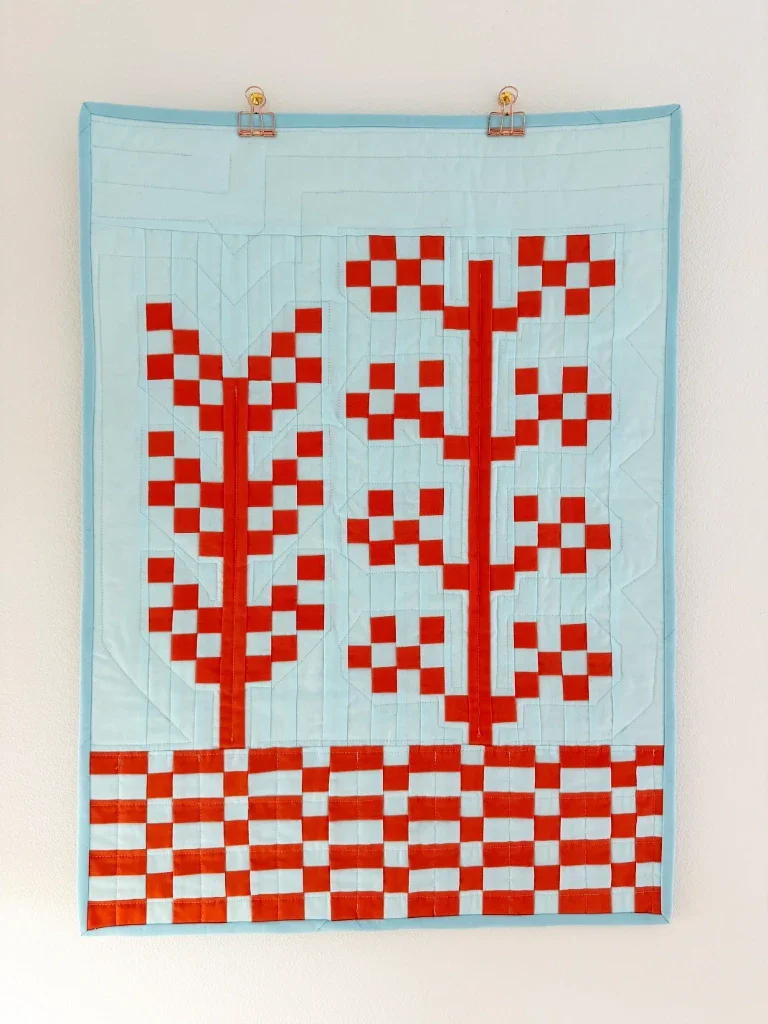 A baby-sized quilt top hanging on a blank off-white wall. The foreground includes 2 pixelated plants in a saturated dark orange, and the background is a very light teal blue. The bottom of the quilt shows a checkerboard pattern in the two colors. Around the quilt is binding in a slightly darker pale teal blue.
