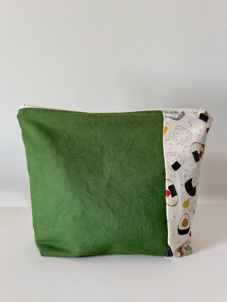 A zippered pouch with a cream zipper and a front panel done in a medium-dark green canvas, along with a strip of heavyweight cream cotton. On the cotton fabric are illustrations of hamsters that are shaped like onigiri and which are holding things like nori strips, shiso leaves, and little octopuses.