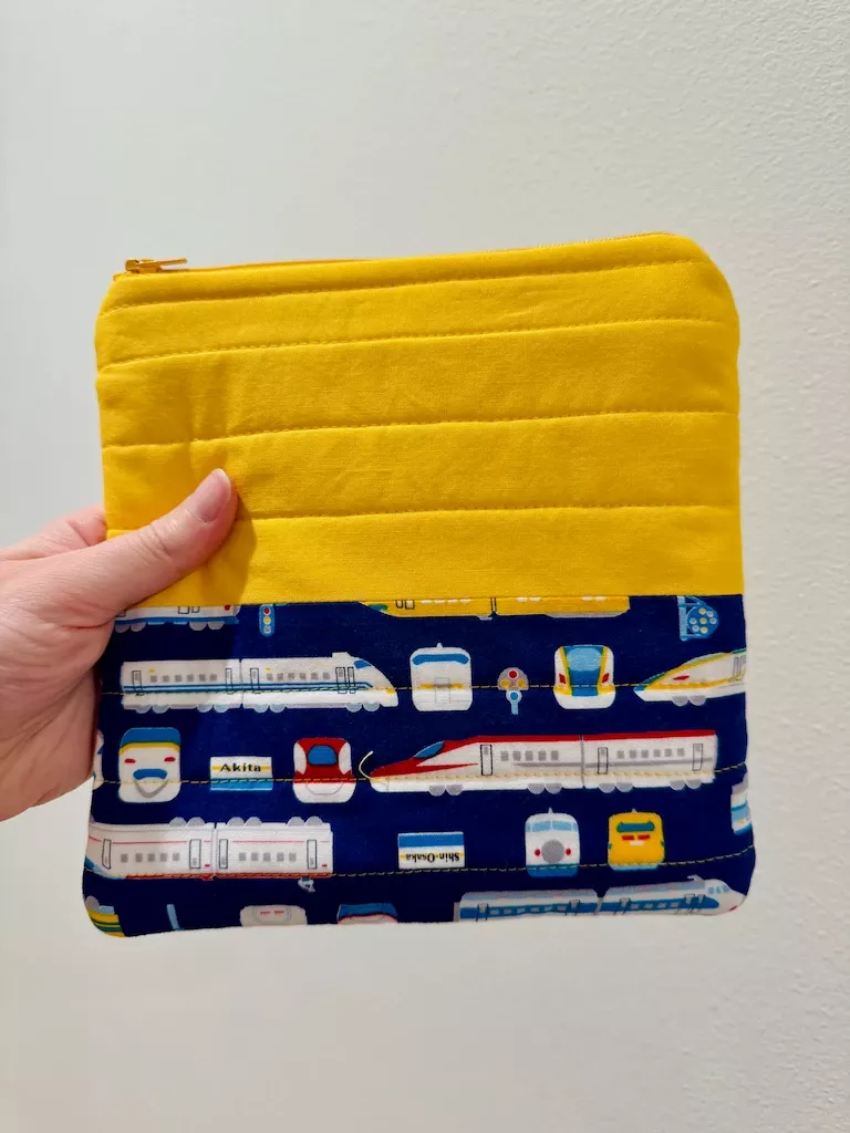 My hand holding an 8 inch square quilted pouch. The top half of the square is a bright saffron yellow, and the bottom is a navy cotton with a printed pattern of Japanese bullet trains in a simple illustrated style