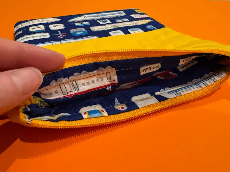 My hand peeling back the top of the pouch to show that the lining is also made from the shinkansen fabric, against a bright orange background