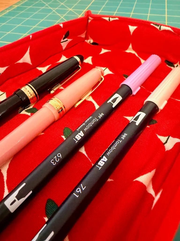 A shallow, rectangular pen tray made of the apple fabric and some coordinating red cotton. Inside the tray are two fountain pens and two Tombow dual brush markers