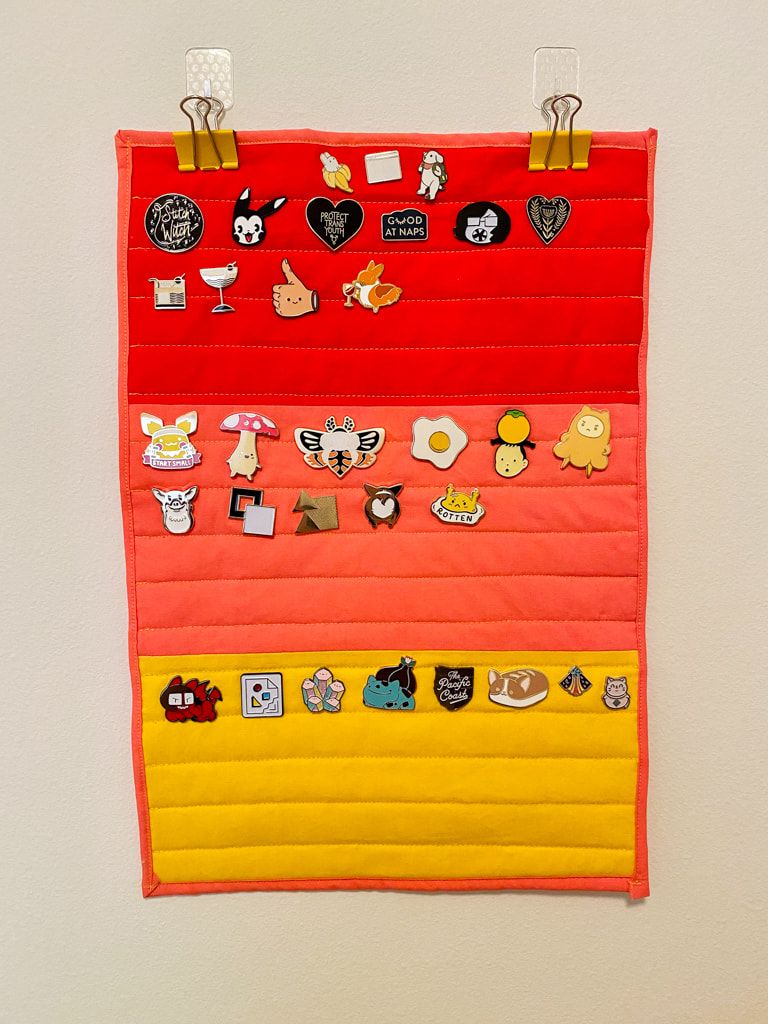 The wall hanging is filled with all manner of cute enamel pins, including 3 corgis, web-themed icons, a couple cocktail, 'protect trans youth', and a couple eggs.