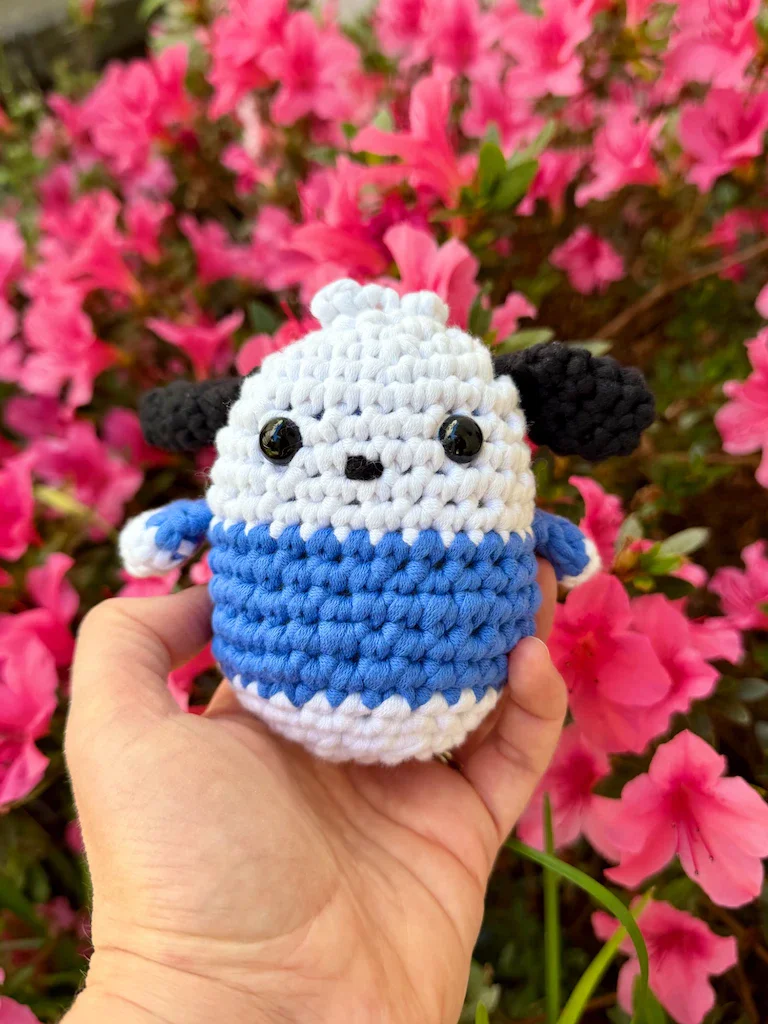 My hand holding an egg-shaped amigurumi plushie of the Sanrio character Pochacco. He is a white dog with black ears and a blue shirt. In the background is a small rhododendron bush in full pink bloom.
