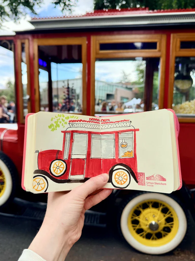 My hand holding up a small, square sketchbook with a drawing of a 1931 popcorn truck on it; the reference vehicle is in the background of the shot. This vintage truck is red, with yellow and white wheels, and has a window in the back showing a popcorn machine. Think of an ice cream truck, but for selling popcorn.