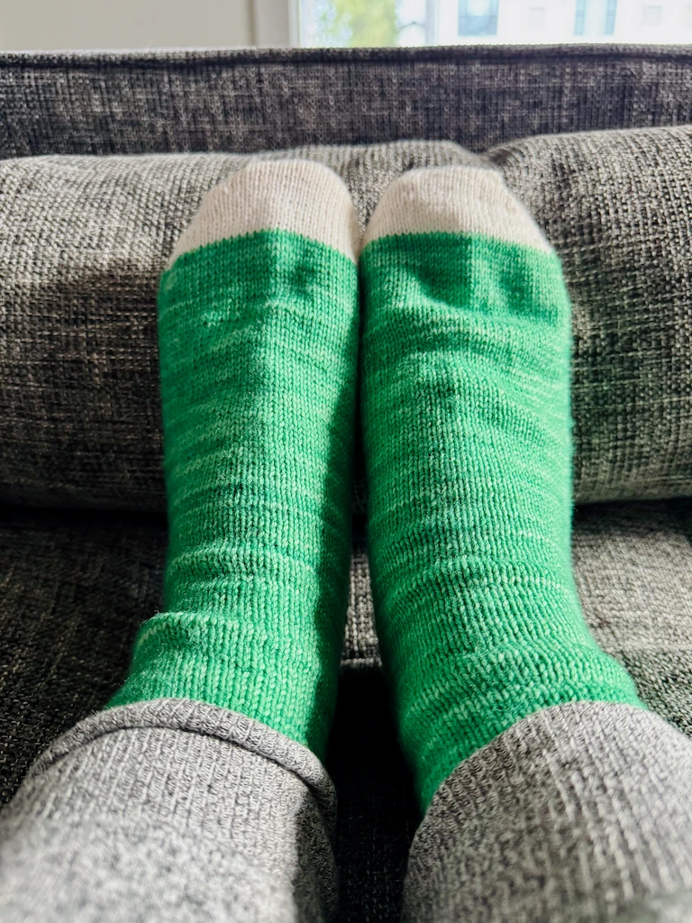 My feet propped up together on a grey couch, clad in home-knitted socks. The socks have light cream toes and a striated medium-teal body.