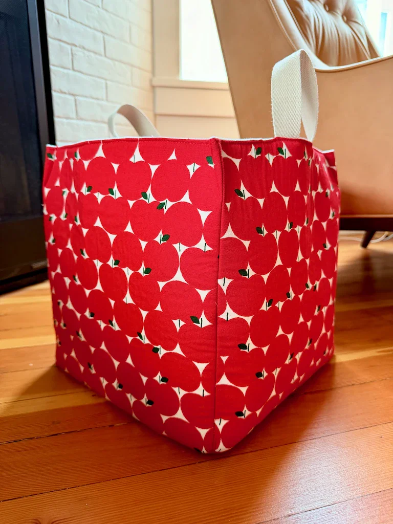 A cotton cube made from a fabric with bright red graphic apples patterned in rows on a white background. There's natural-colored handles poking out the top of two sides. The cube is sitting on a warm wood floor.